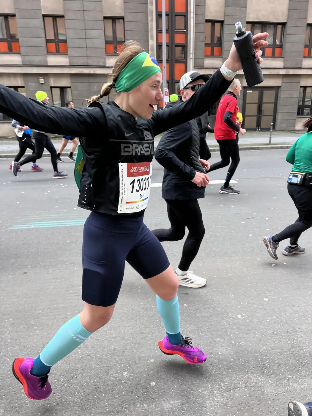 Zoe running during the Berlin half marathon, arms wide, bib 13033, Brazil headband, blue socks